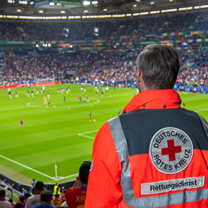 Sanitäter des Deutschen Roten Kreuzes in orange-blauer Einsatzkleidung steht im Stadion und blickt aufs Spielfeld.